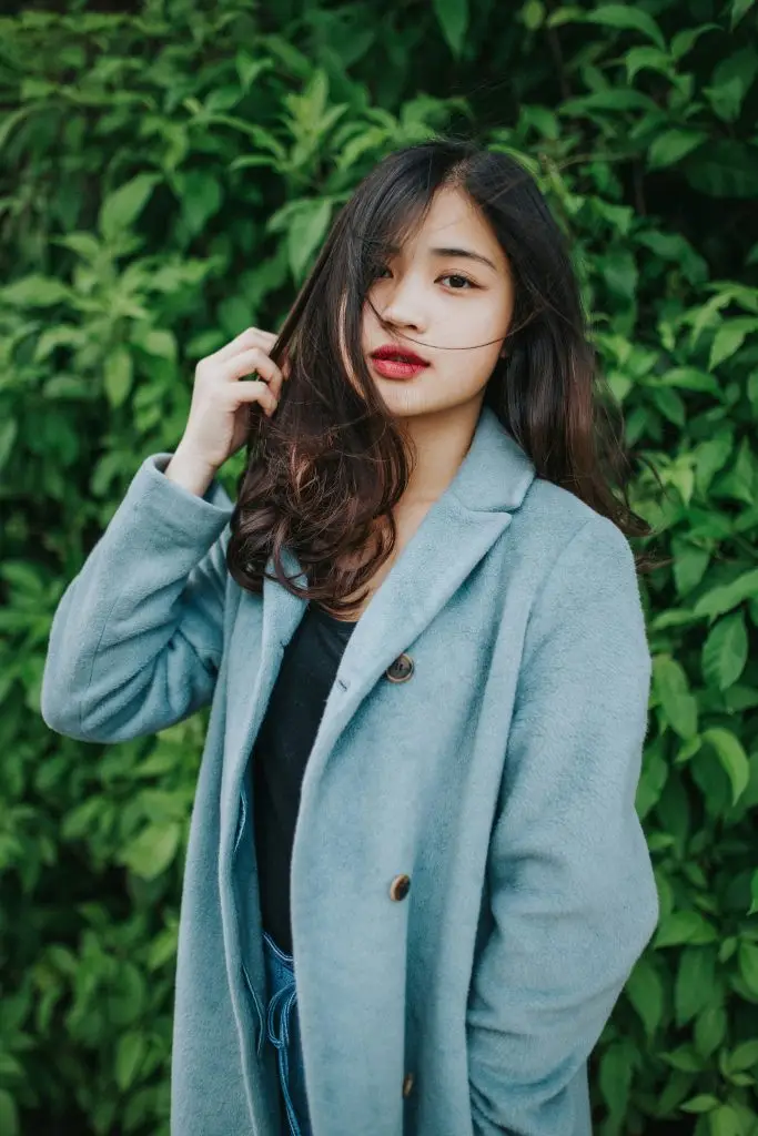 Petite woman wearing a light blue tailored coat over a black top, standing outdoors against lush greenery, highlighting a sleek and flattering petite outerwear fit.