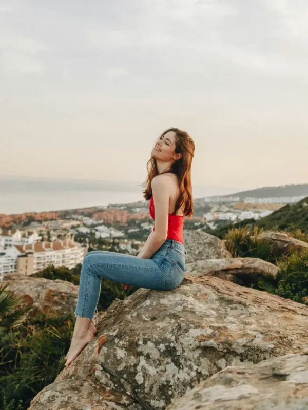 Petite woman wearing fitted blue jeans and a red sleeveless top, seated on a rocky overlook with a coastal city view, showcasing a flattering petite fit suitable for everyday wear.
