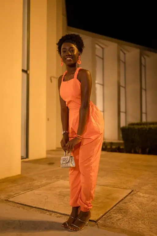 A petite woman in a orange fashionable jumpsuit.