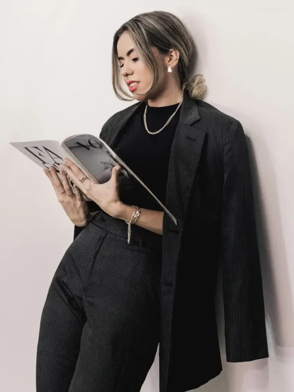 Petite woman wearing tailored black pinstripe pants with a matching blazer and black top, standing indoors while reading a magazine, showcasing a polished and professional petite fit.