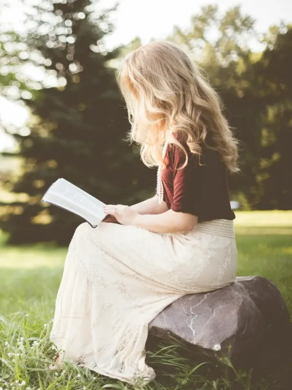 Petite woman wearing a flowing light-colored skirt and short-sleeve top, seated on a rock while reading outdoors in a sunlit grassy setting, highlighting an elegant and comfortable petite skirt fit.